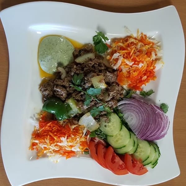 Indian keema with green sauce, shredded carrots, red onions, cucumber, and tomatoes on a white plate.