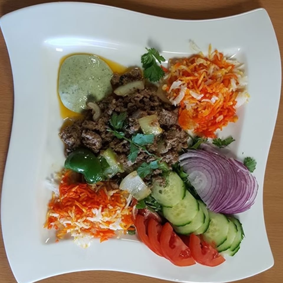 Indian keema with green sauce, shredded carrots, red onions, cucumber, and tomatoes on a white plate.