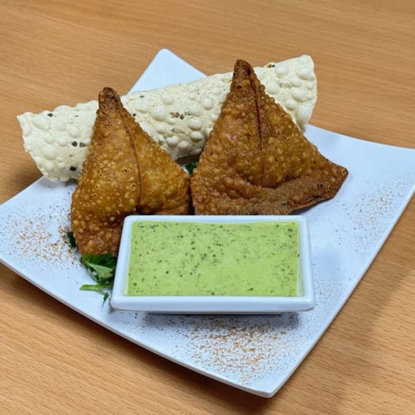 Two golden-brown samosas and papadum on a white plate with a side of green chutney.