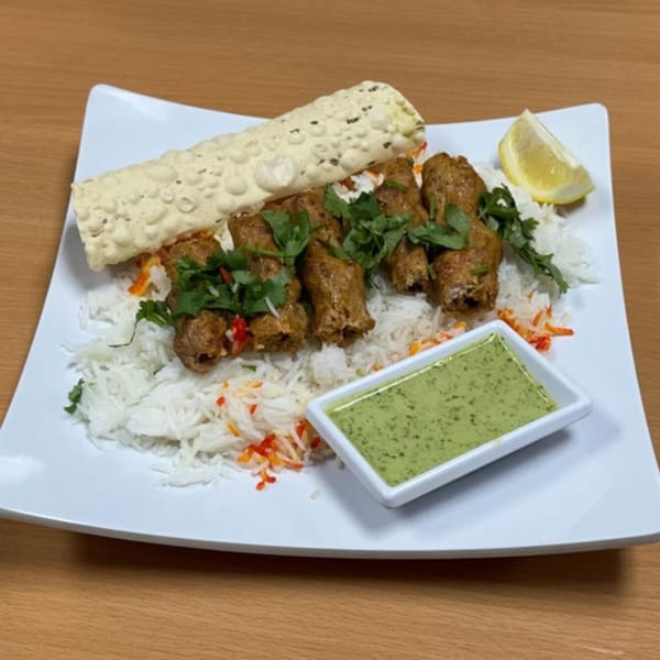 Indian seekh kebab with rice, papadum, green chutney, and a lemon wedge on a white plate.