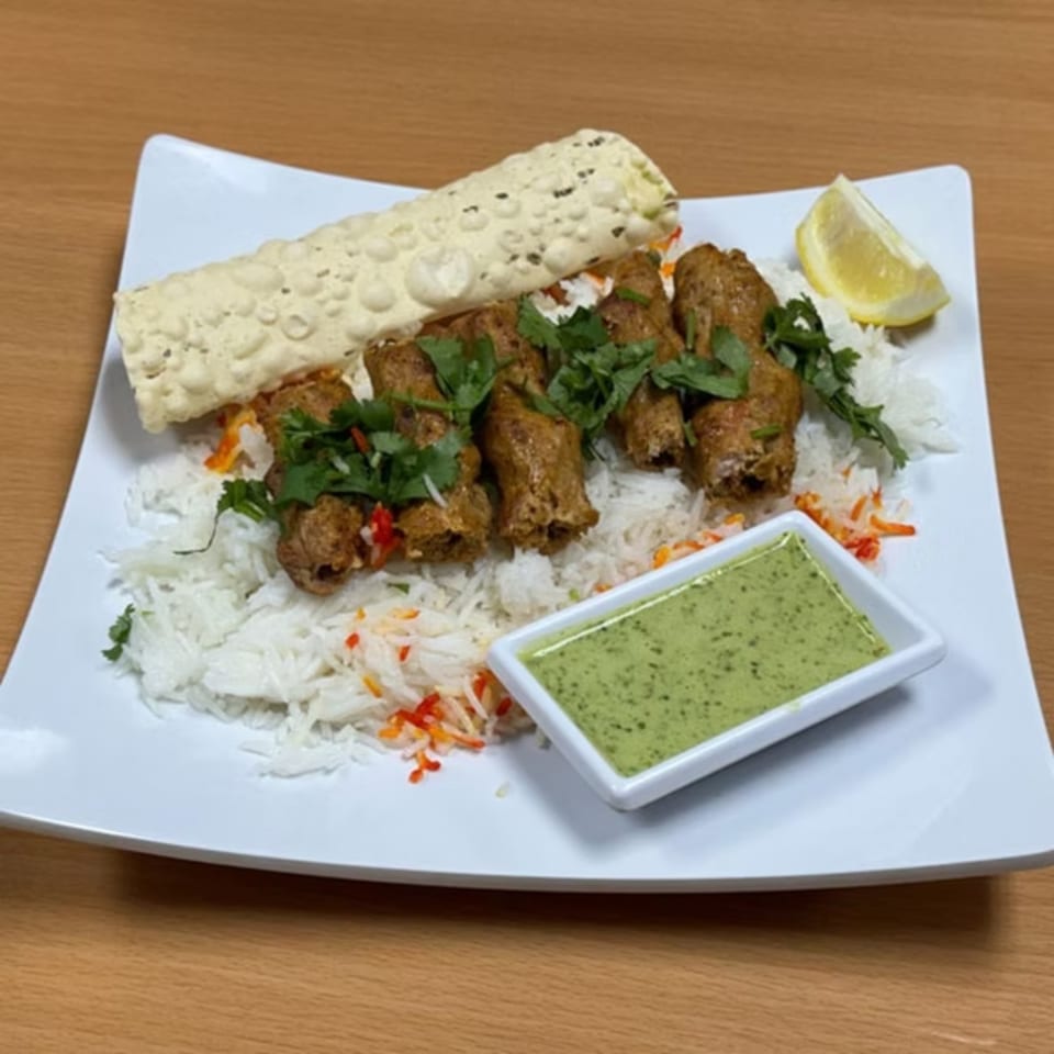 Indian seekh kebab with rice, papadum, green chutney, and a lemon wedge on a white plate.
