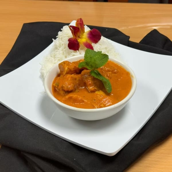A bowl of orange chicken curry with a mint garnish, served with white rice and an orchid.