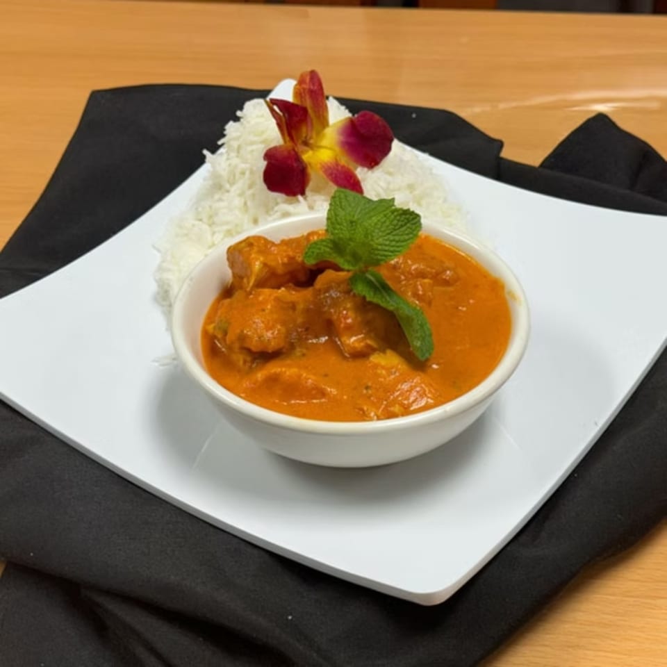 A bowl of orange chicken curry with a mint garnish, served with white rice and an orchid.