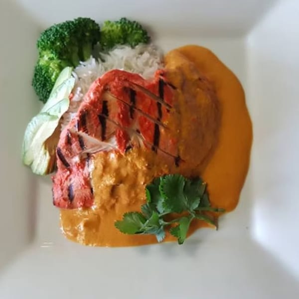 Grilled red chicken tikka with orange curry sauce, white rice, broccoli, and cilantro on a white plate.